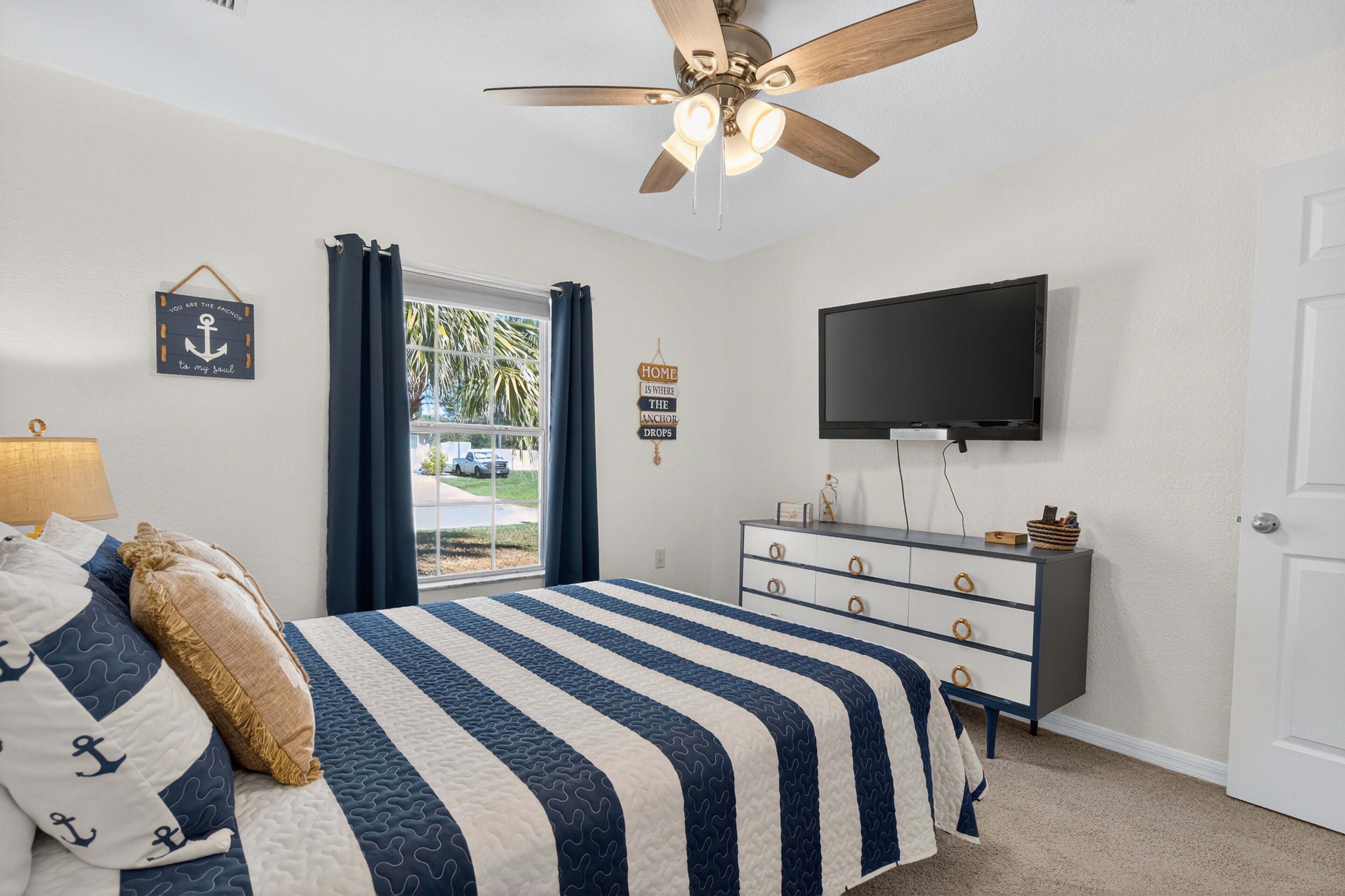 A bedroom with a bed, a ceiling fan, a television, a dresser, and a window with curtains.