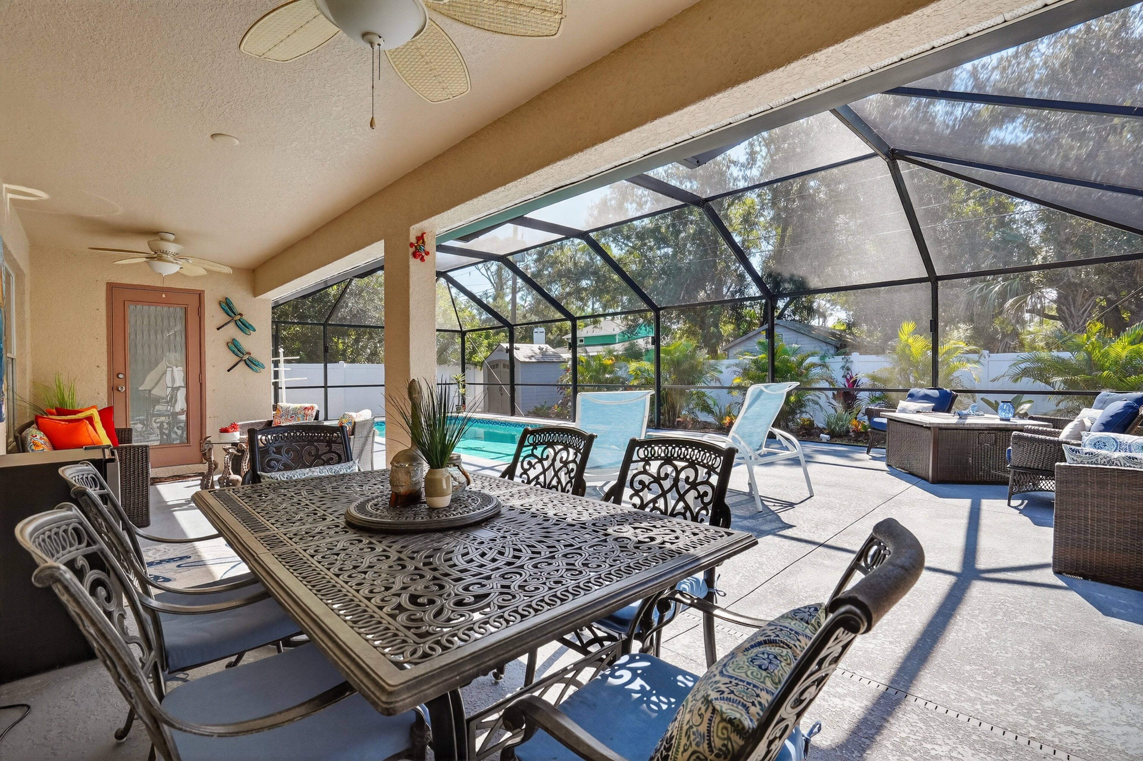 An outdoor patio with a table and chairs, a ceiling fan, and a view of a swimming pool and greenery.