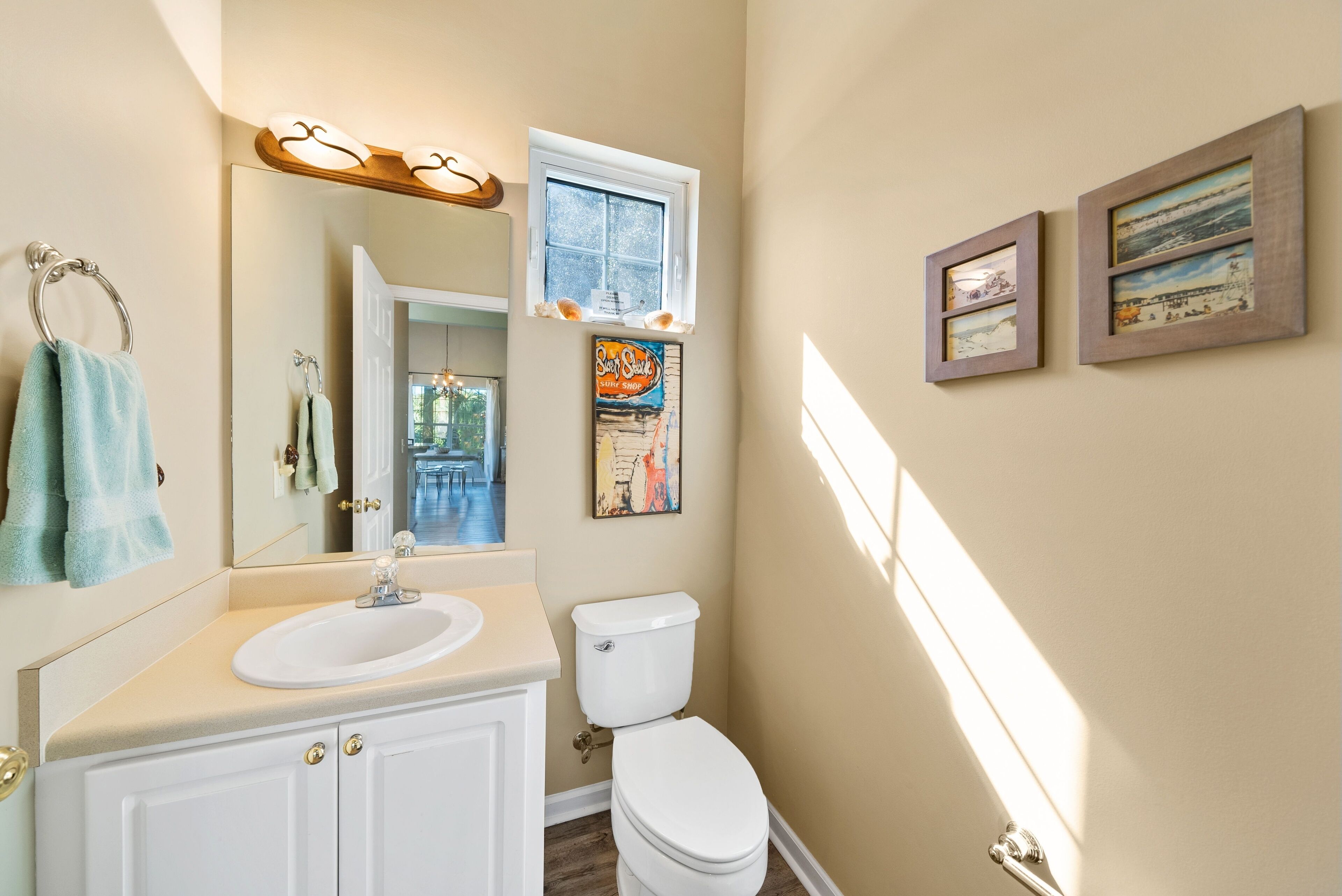 Une salle de bain avec un lavabo blanc, des toilettes et une fenêtre située au-dessus des toilettes. Deux tableaux encadrés sont accrochés au mur.