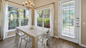 A dining area with a wooden table, clear chairs, a chandelier, and large windows with curtains. - Blue Palm 3 Bedroom Home (Panama City Beach)