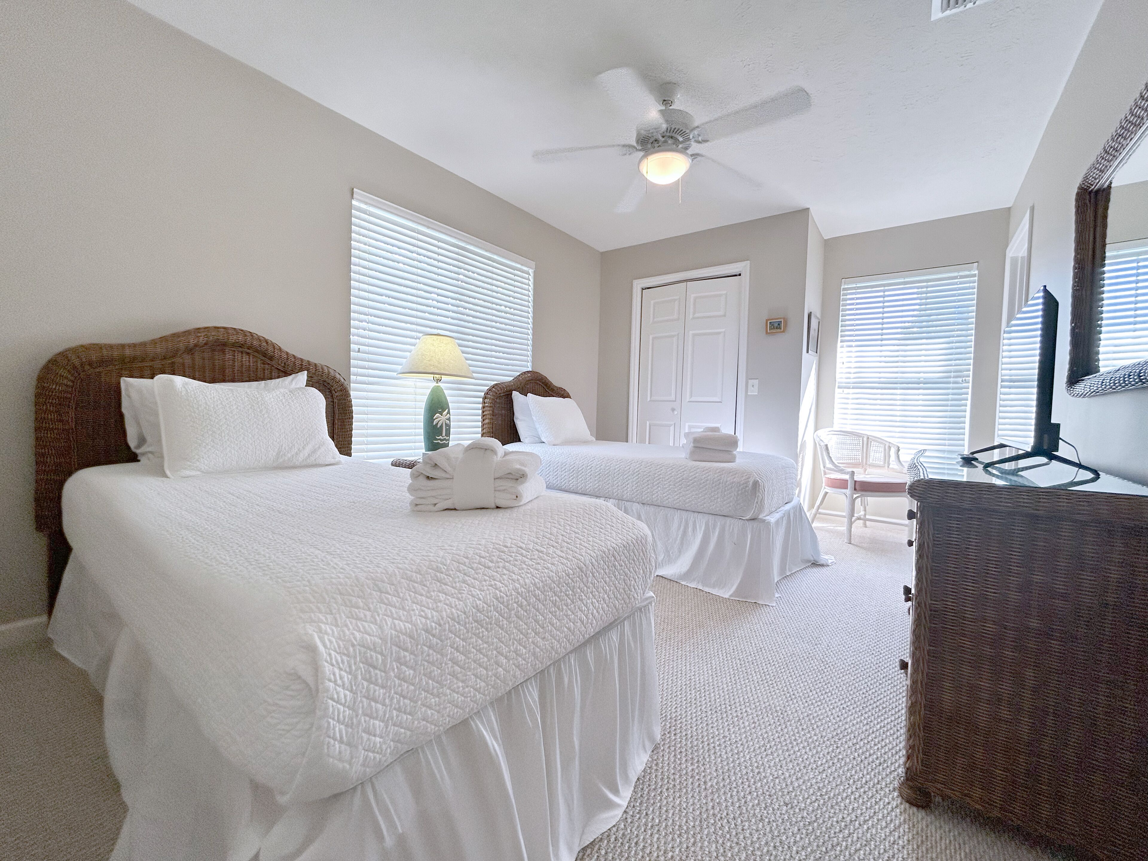 A hotel room with two beds, a ceiling fan, a lamp, and a wicker chair.