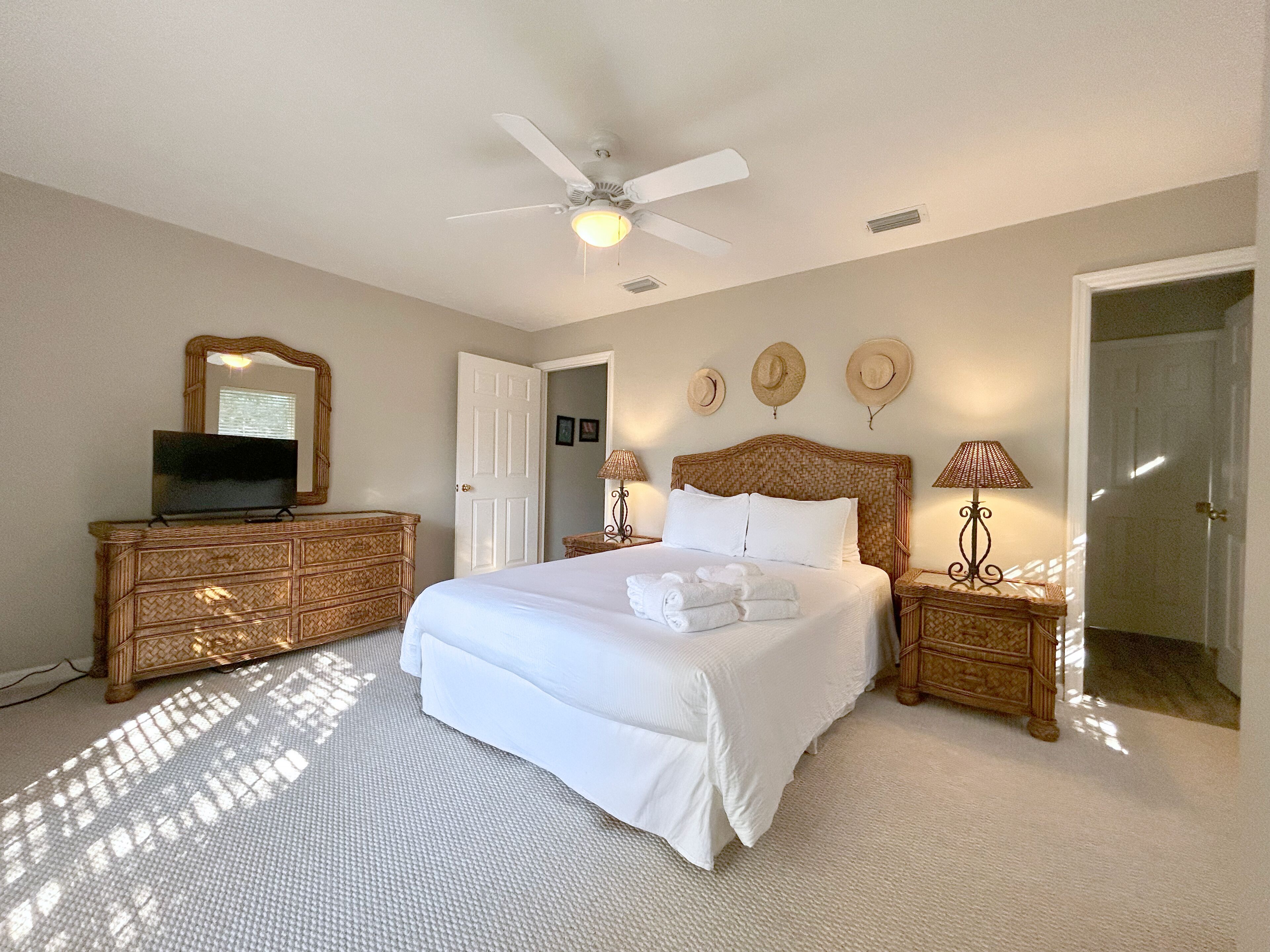 Une chambre avec un grand lit, un ventilateur de plafond, un téléviseur posé sur une commode et trois chapeaux accrochés au mur.
