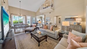 A spacious living room with a high ceiling, a balcony, and a large TV. - Blue Palm 3 Bedroom Home (Panama City Beach)