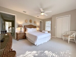 A bedroom with a large bed, two bedside tables with lamps, a ceiling fan, a chair, and a door. - Blue Palm 3 Bedroom Home (Panama City Beach)
