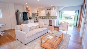 Una cocina moderna con armarios blancos, un refrigerador de acero inoxidable y un microondas. Hay un comedor con una mesa de vidrio y un sofá.