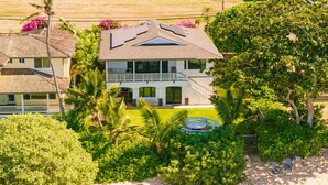 Una casa de dos pisos con balcón, rodeada de abundante vegetación y una playa arenosa.