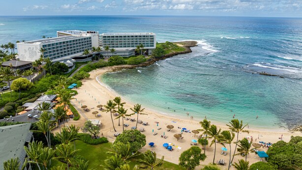 Un hotel frente a la playa con varias edificaciones, una playa de arena y aguas azules cristalinas.