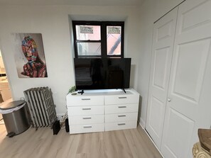 Una habitación con un aparador blanco, un radiador, un cesto de basura y una ventana que ofrece vistas a edificios.