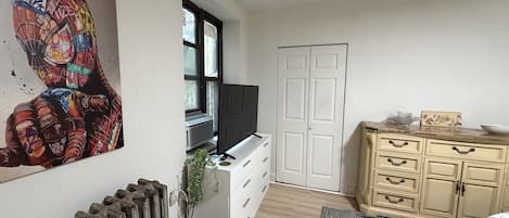 Una habitación con un radiador, un televisor sobre un mueble bajo, un armario blanco y un cuadro en la pared.