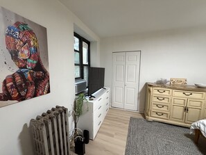 Una habitación con un radiador, un televisor sobre un mueble bajo, un armario blanco y un cuadro en la pared.