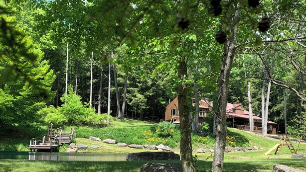 A lakeside cabin with a dock, surrounded by dense forest and a grassy area with a playground. - This beautiful Lodge in the woods is perfect for year round enjoyment! (Allegany)