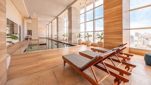 A modern indoor pool area with lounge chairs and large windows. - Apartamento Moderno Premium - Cittyplex  (Osasco)