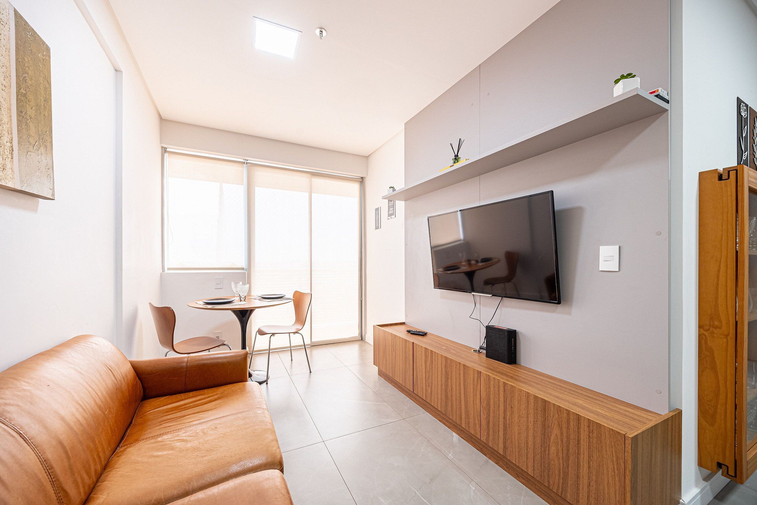 A modern living room with a leather sofa, a TV mounted on the wall, a wooden cabinet, and a dining area with a table and chairs.