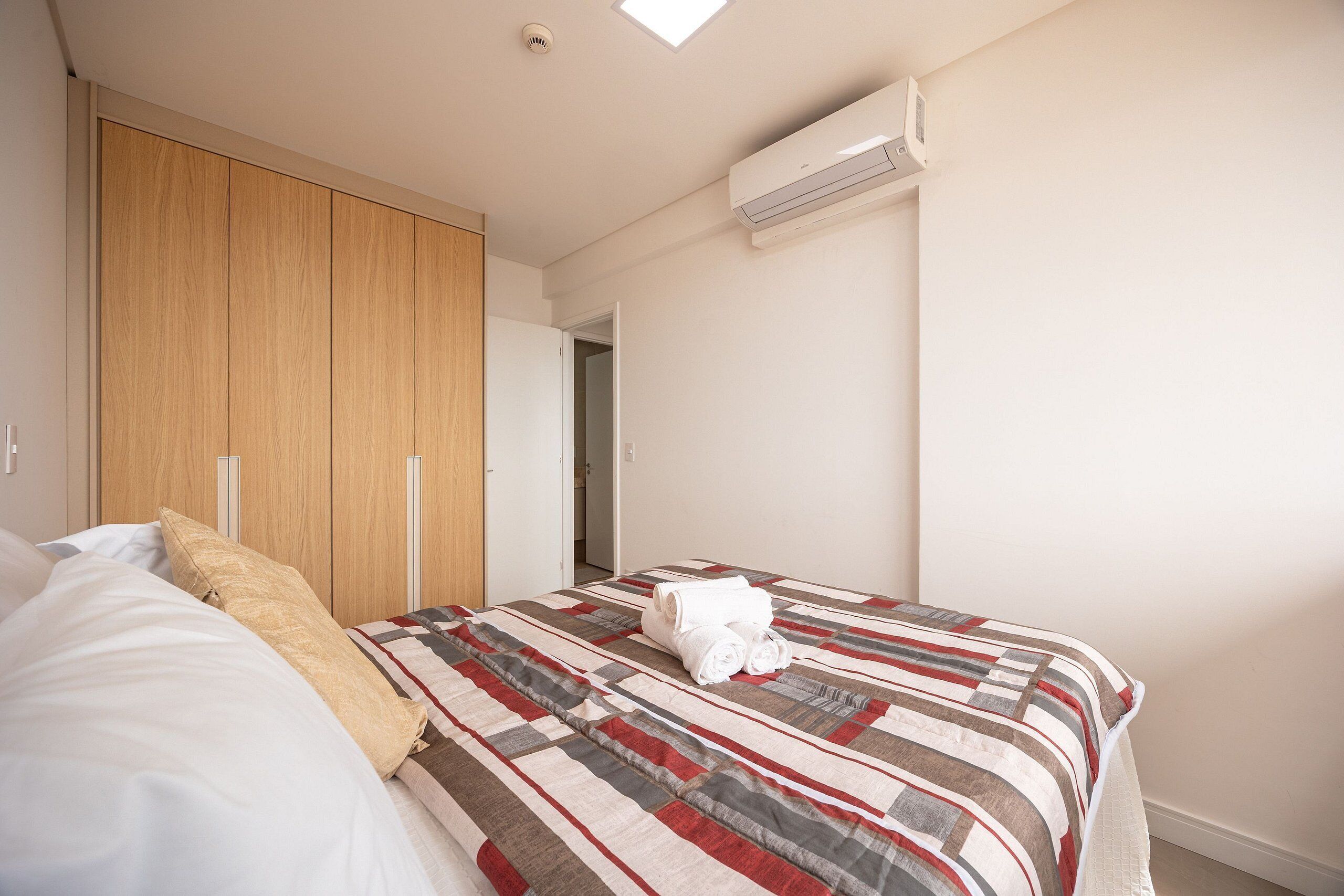 A bedroom with a bed, a wooden wardrobe, an air conditioning unit, and a door leading to another room.