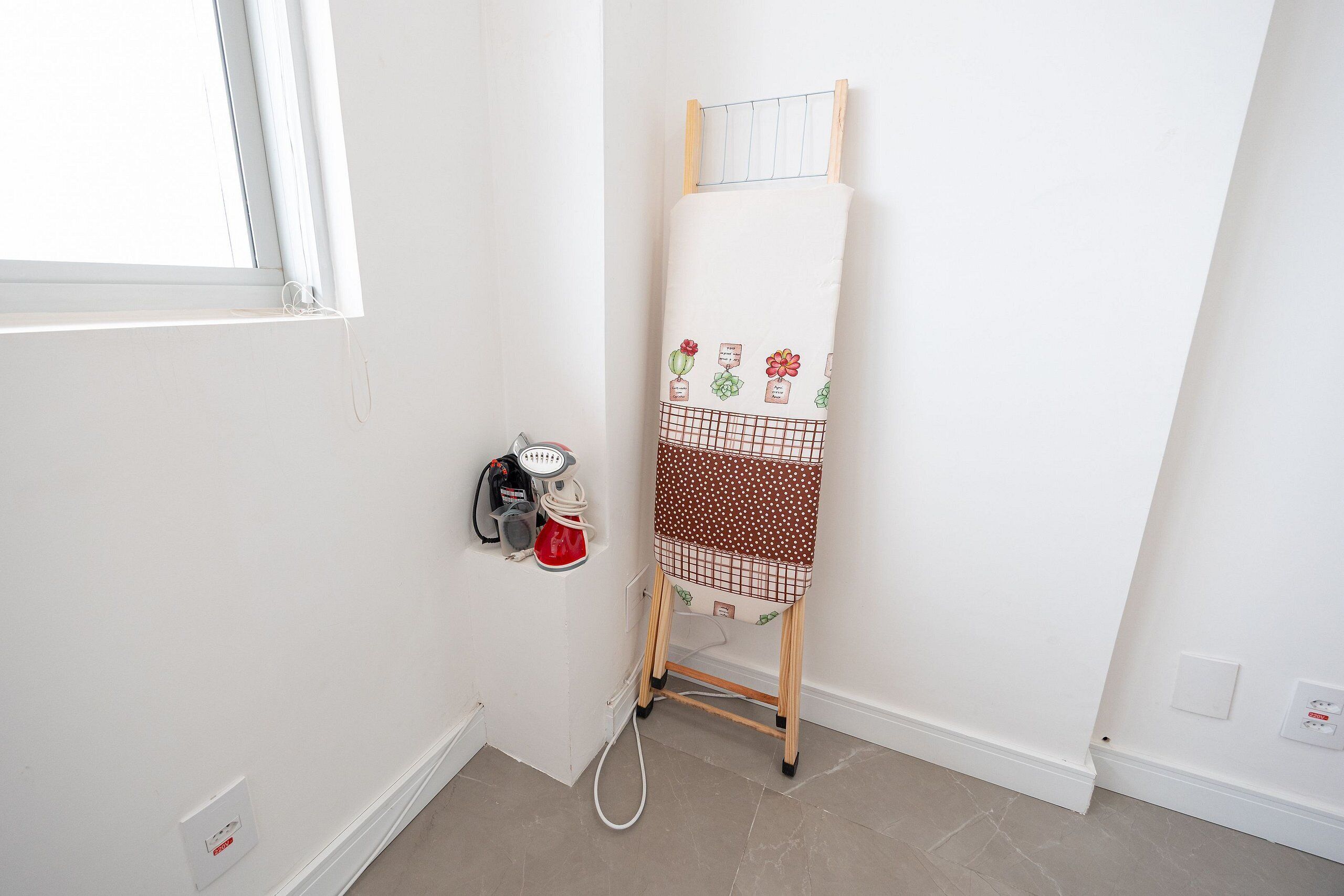 A room with a window, an ironing board, a kettle, and a power outlet.