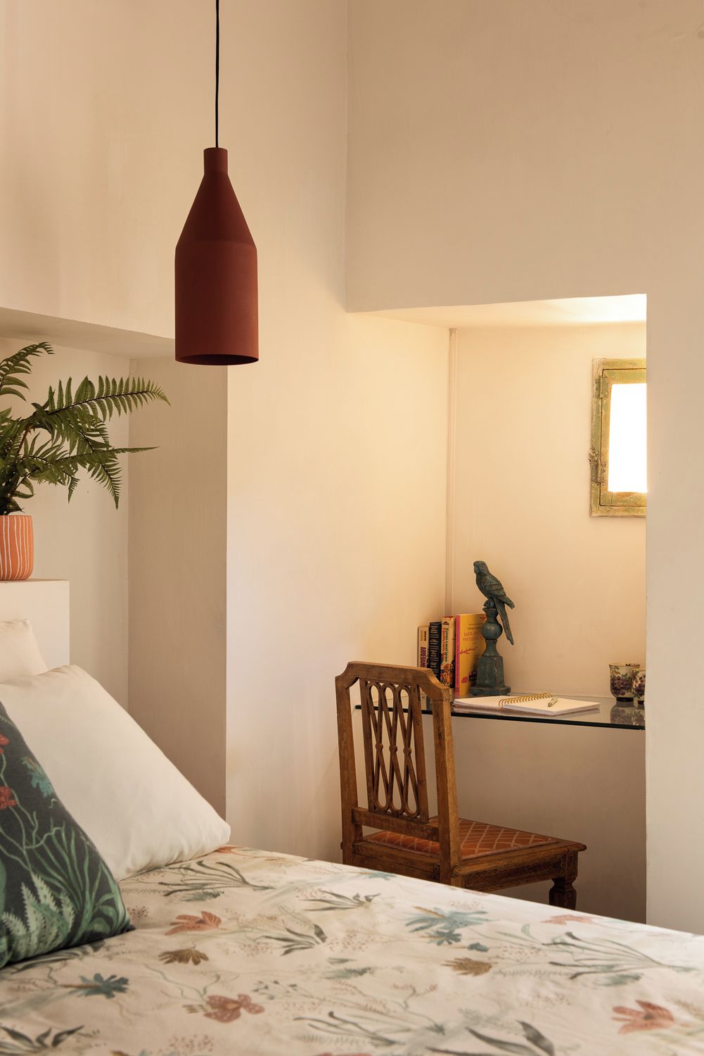 A bedroom with a bed, a chair, a desk, a hanging light, and a plant.