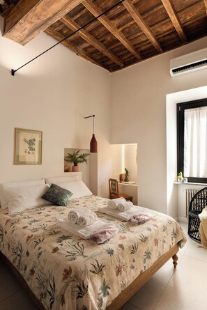 Une chambre avec un plafond en bois, un lit recouvert d’une literie à motifs floraux, une table de chevet avec une lampe et une fenêtre avec des rideaux.