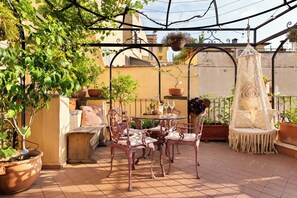 An outdoor patio with a table set for dining, surrounded by potted plants and a hanging chair.