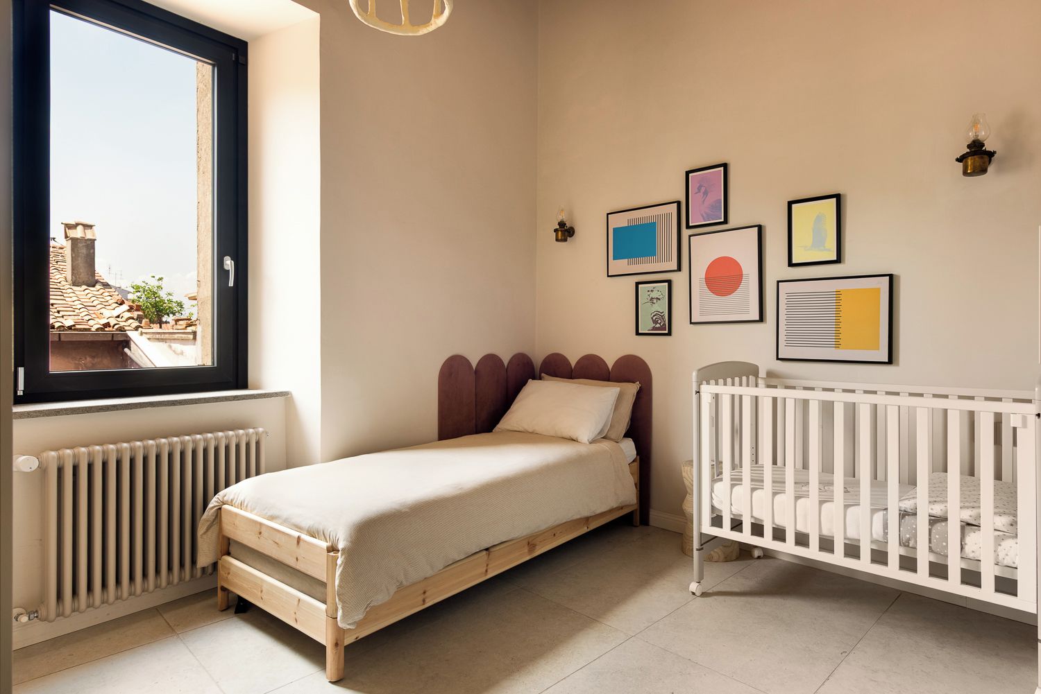 A bedroom with a single bed, a crib, a radiator, a window with a view, and wall art.