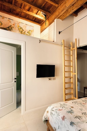 A bedroom with a bed, a television on the wall, a wooden ladder, and a door leading to another room.