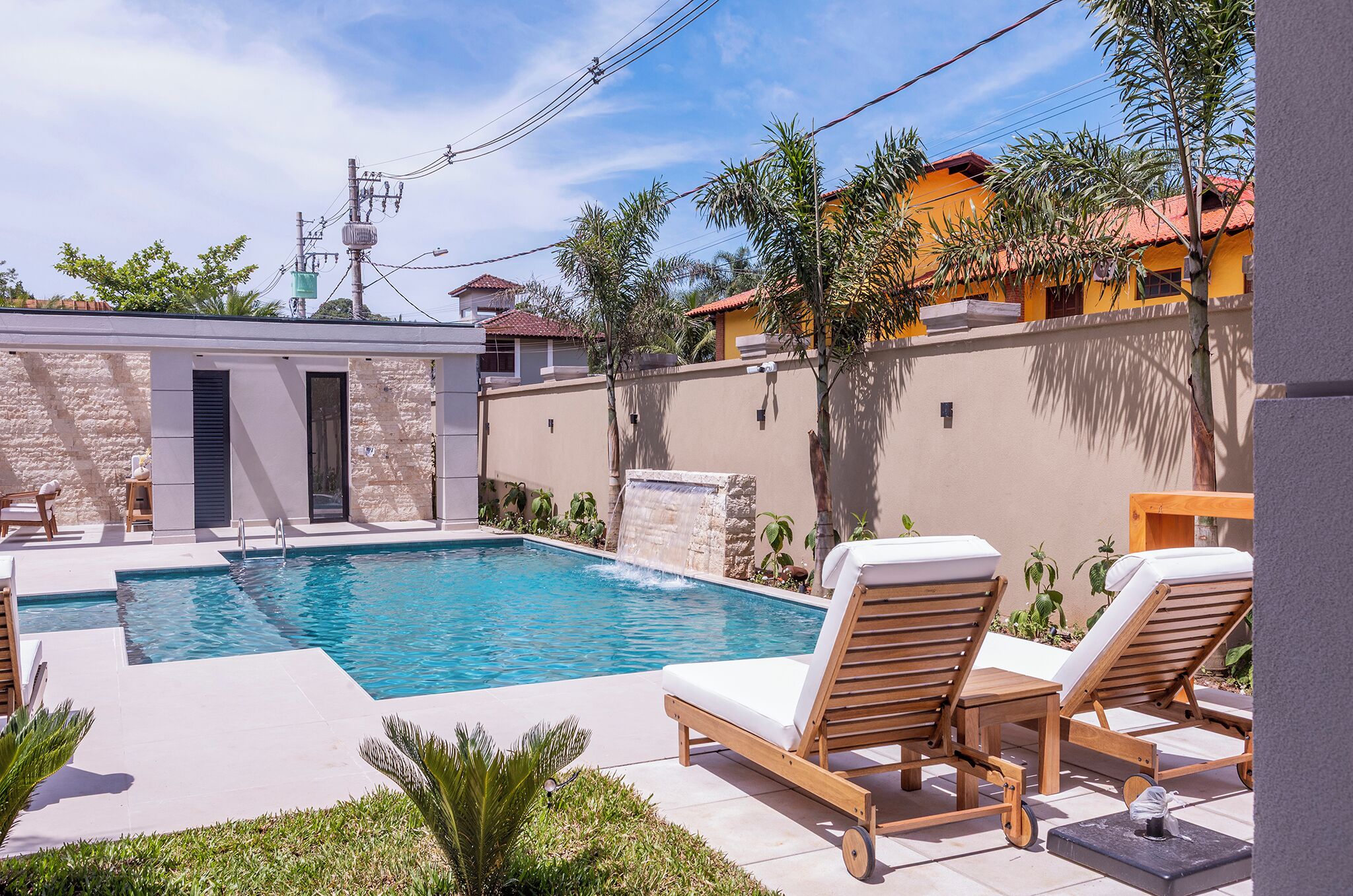 Piscina com espreguiçadeiras, parede de pedra e pequena cascata.