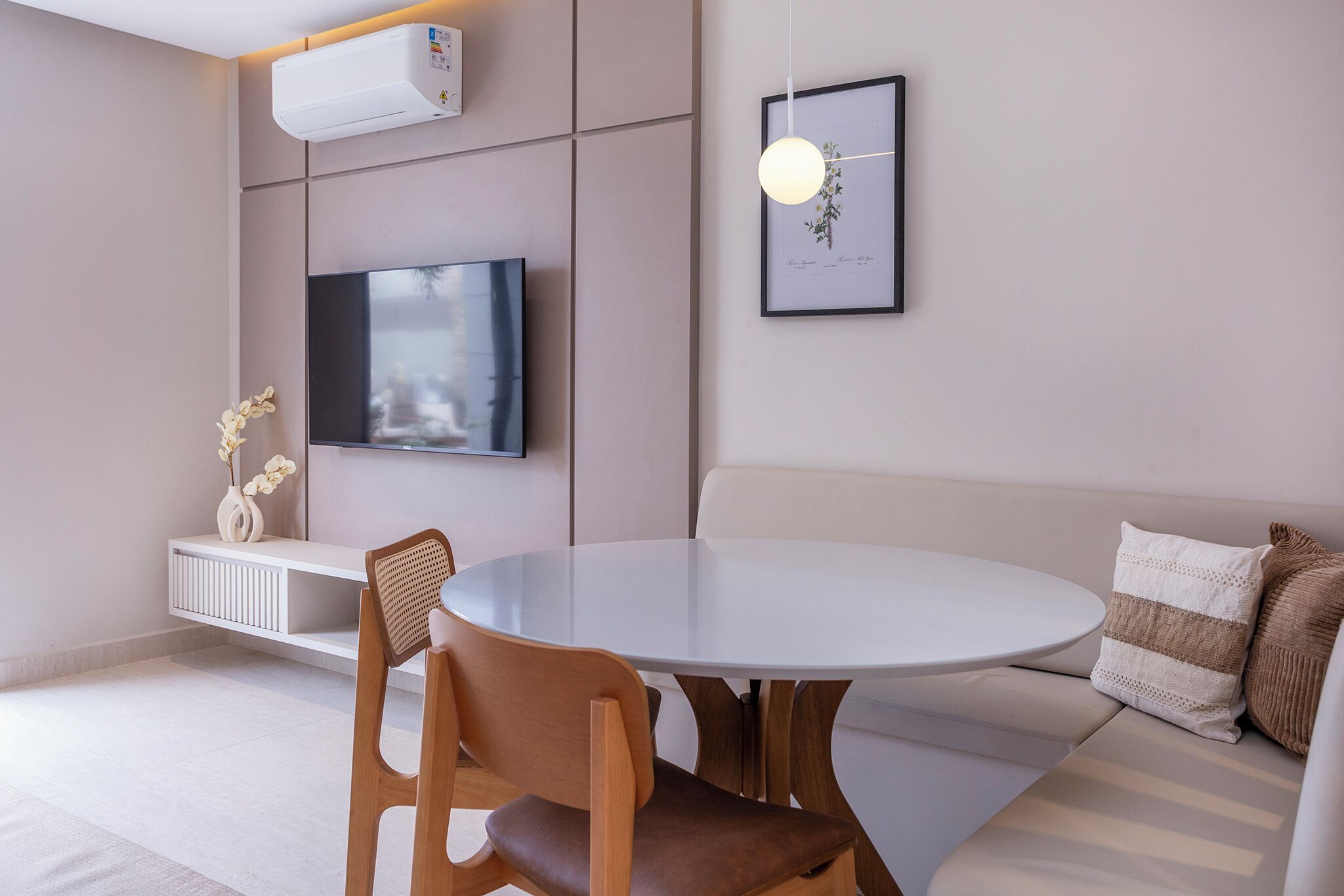 A modern living room with a round white table, a wooden chair, a sofa with striped cushions, a mounted TV, and a wall-mounted air conditioner.