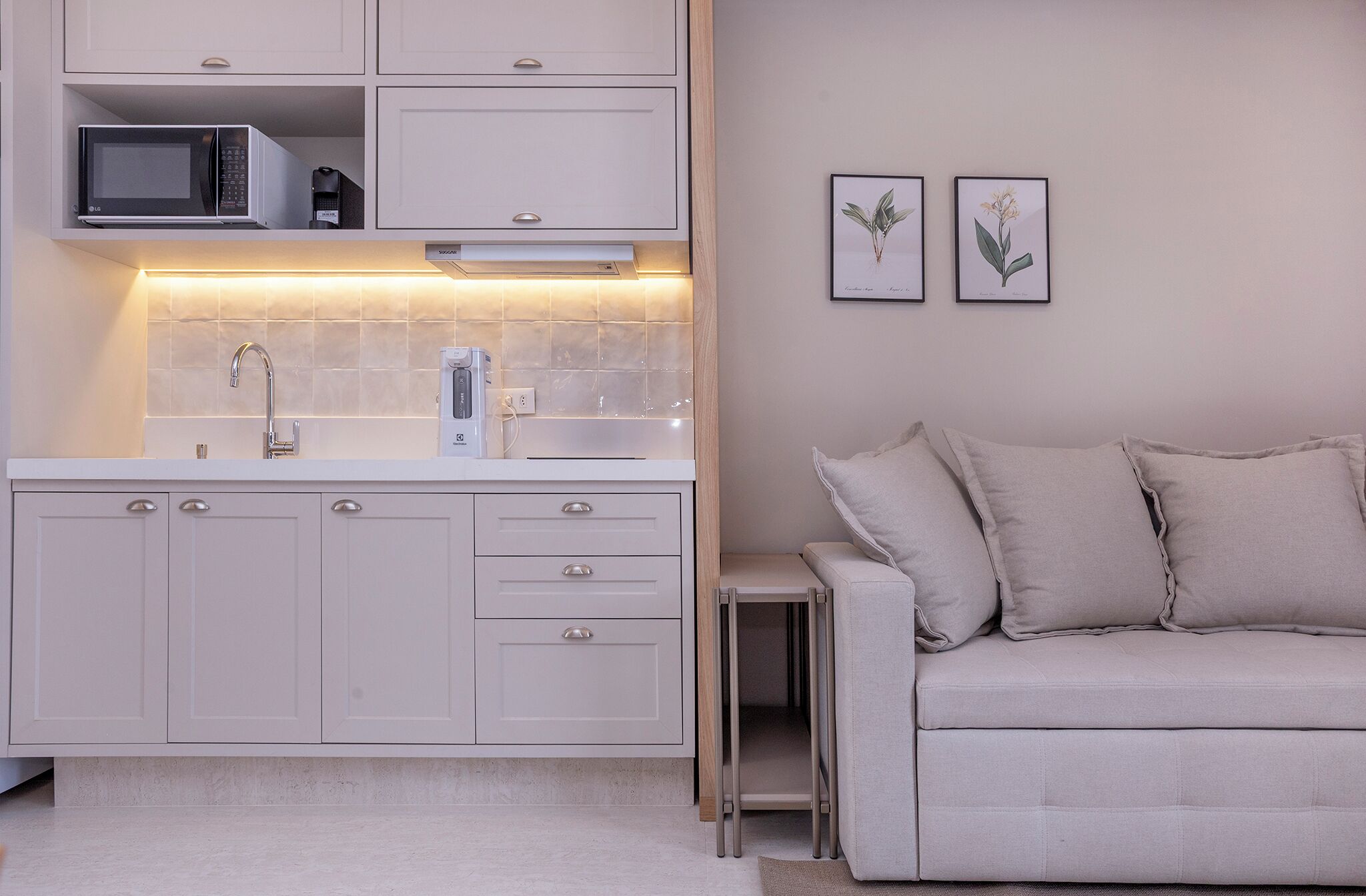 A modern kitchen with white cabinets, a microwave, and a built-in oven. There is a small side table and a grey sofa with cushions. Two framed botanical illustrations are on the wall.