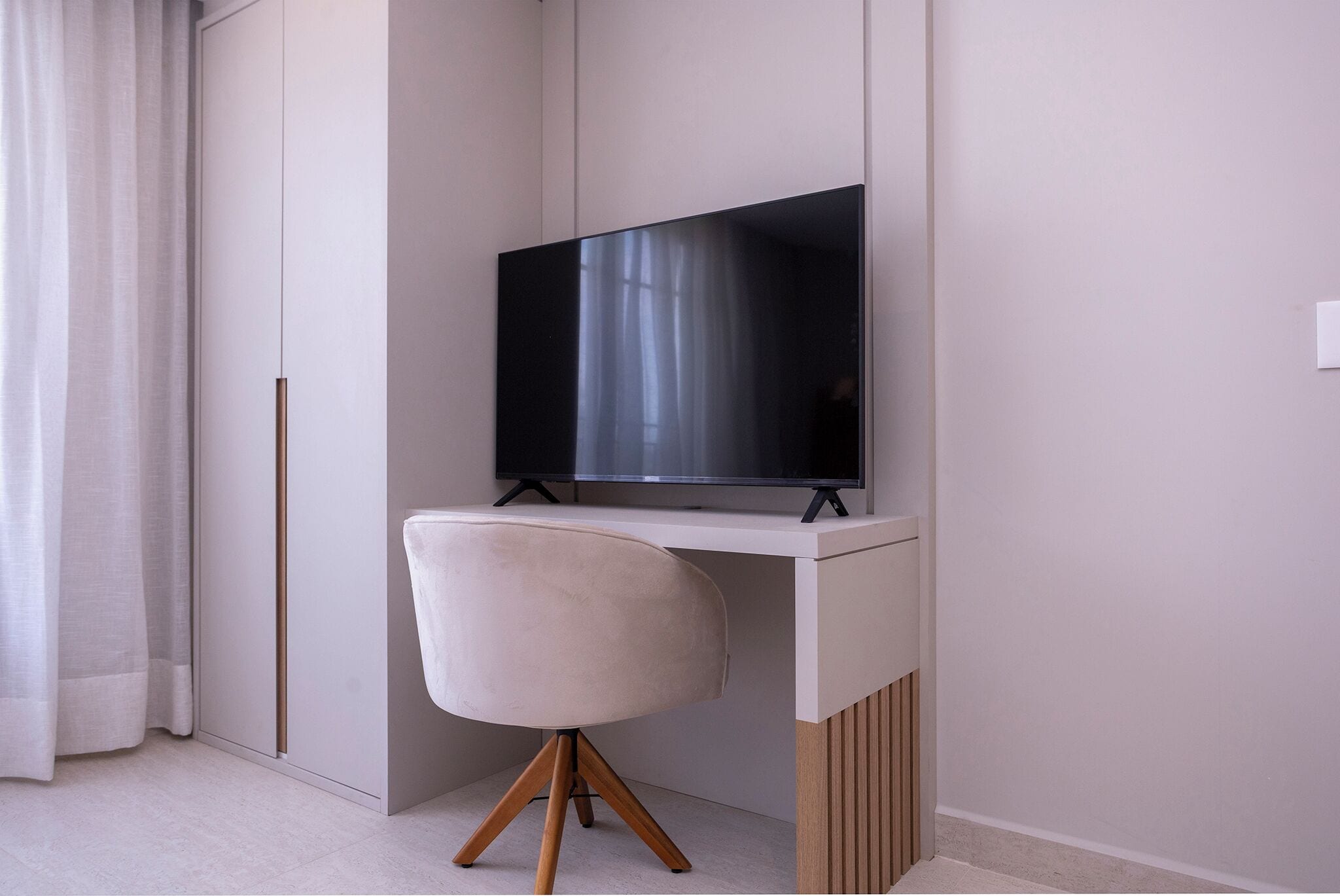A modern room with a flat-screen TV mounted on a white desk, a light-colored chair, and a curtain on the left.
