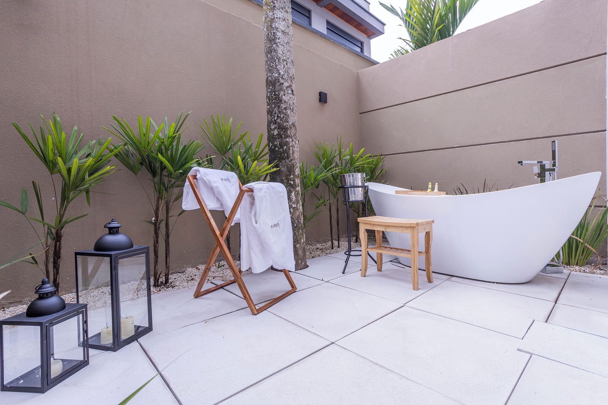 A modern outdoor bathroom with a freestanding bathtub, a wooden stool, and a towel rack.