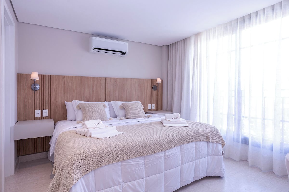 Ein modernes Hotelzimmer mit einem großen Bett, einem hölzernen Kopfteil, zwei Wandleuchten, einer Klimaanlage und transparenten Vorhängen.