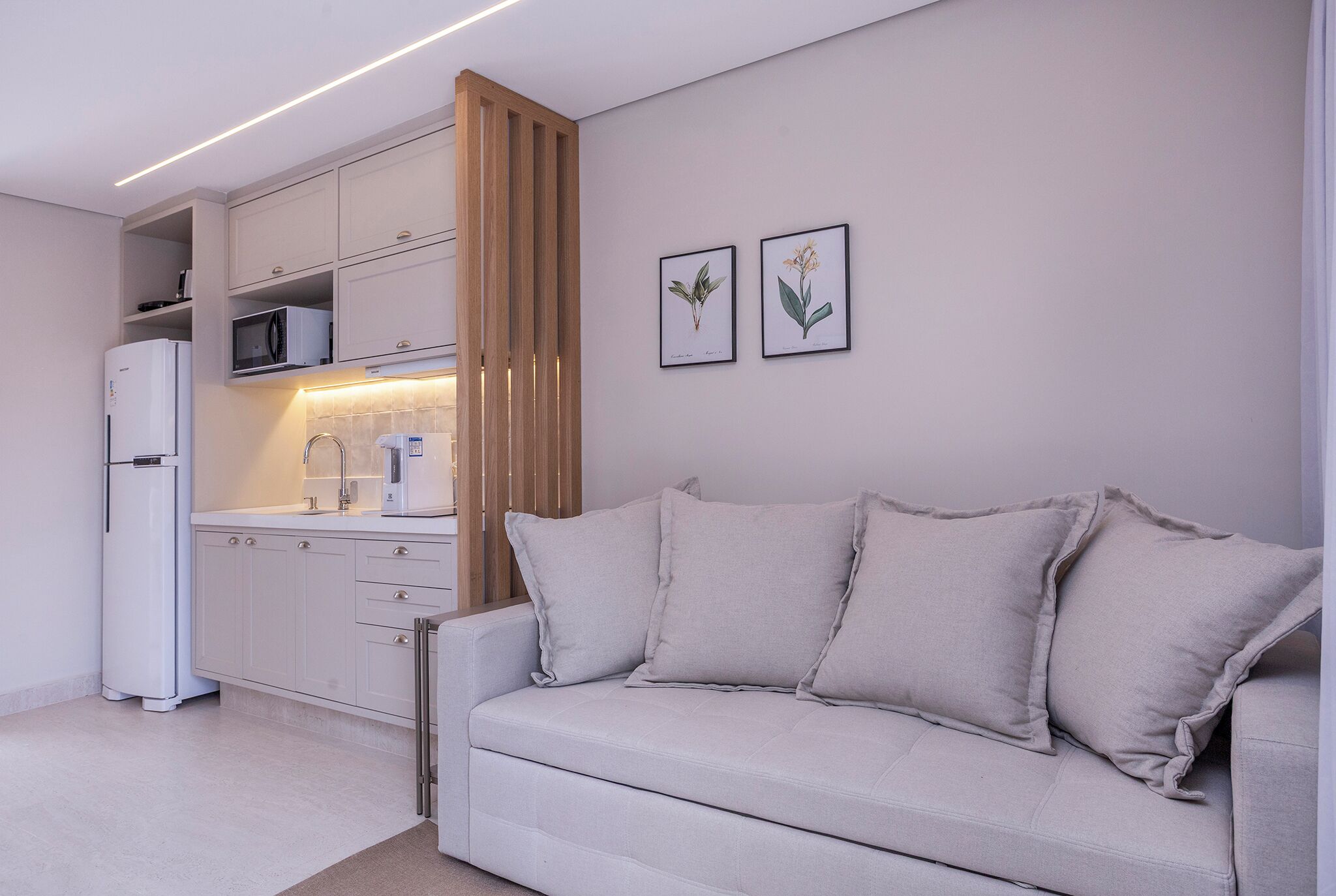 A modern kitchen with white cabinets, a white refrigerator, and a microwave. There is a beige sofa with cushions and two framed botanical prints on the wall.