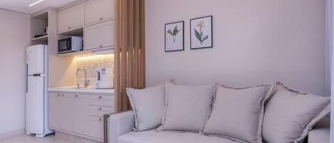 A modern kitchen with white cabinets, a white refrigerator, and a microwave. There is a beige sofa with cushions and two framed botanical prints on the wall.