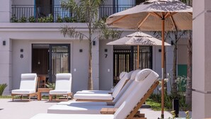 A poolside area with lounge chairs and umbrellas, a wooden table, and a building with numbered doors. - Solena Hotel (São Sebastião)