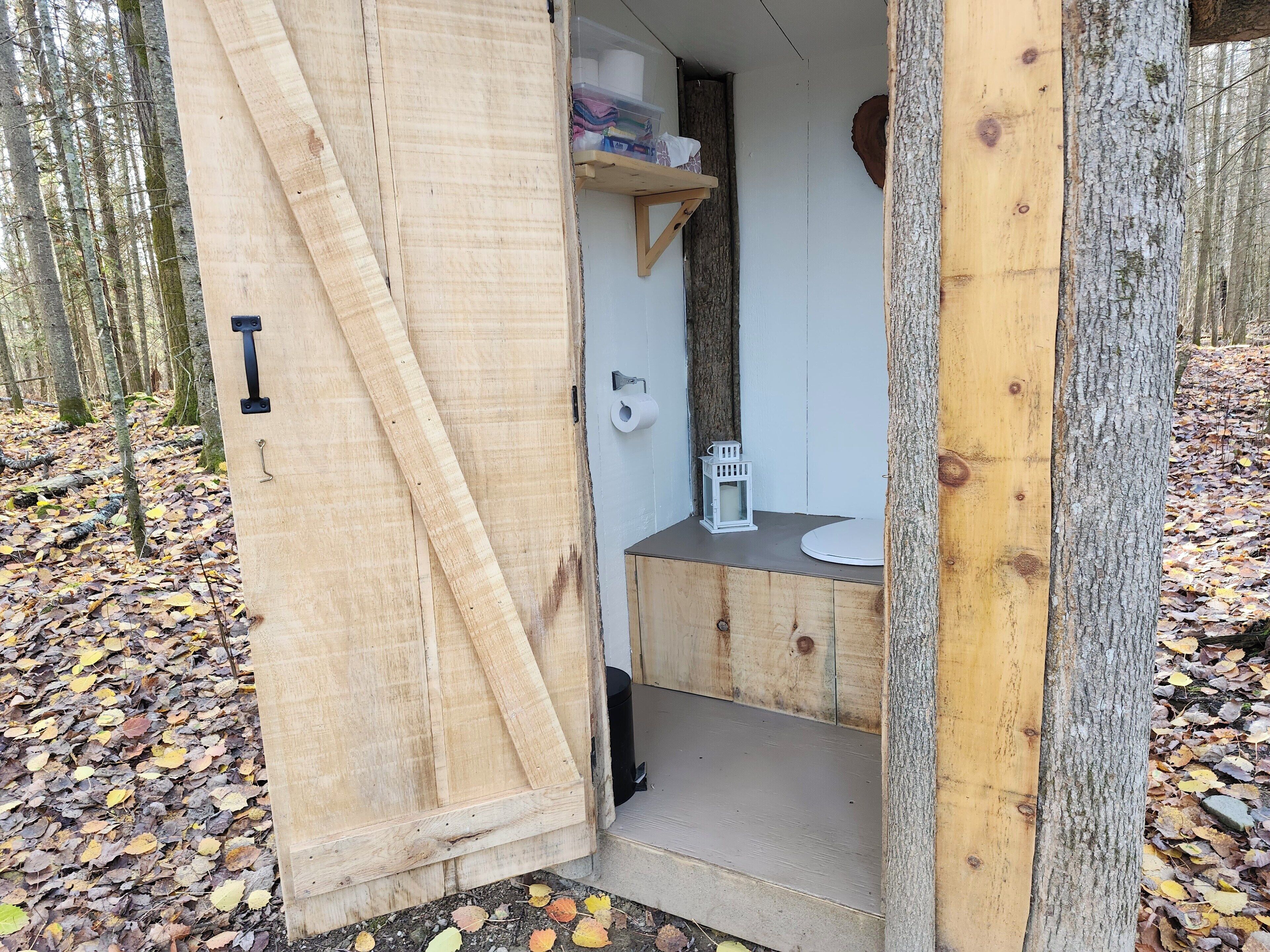 A wooden cabin with a door open, revealing a small bathroom with a toilet, sink, and shelves with toiletries.