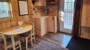 A wooden interior with a round table, chairs, a small kitchen area, and a door leading outside. - The Rustic Treehouse Hideaway-cozy treehouse retreat in Northern Ontario (Corbeil)
