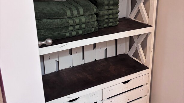 A white wooden shelf unit with three drawers and two shelves, each holding neatly folded green towels.