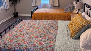 A bedroom with a large bed, a smaller bed, a ceiling fan, a window with curtains, a potted plant, and a nightstand with a lamp.