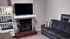 A modern living room with a flat-screen TV, a fireplace, a ceiling fan, and a gray sofa.
