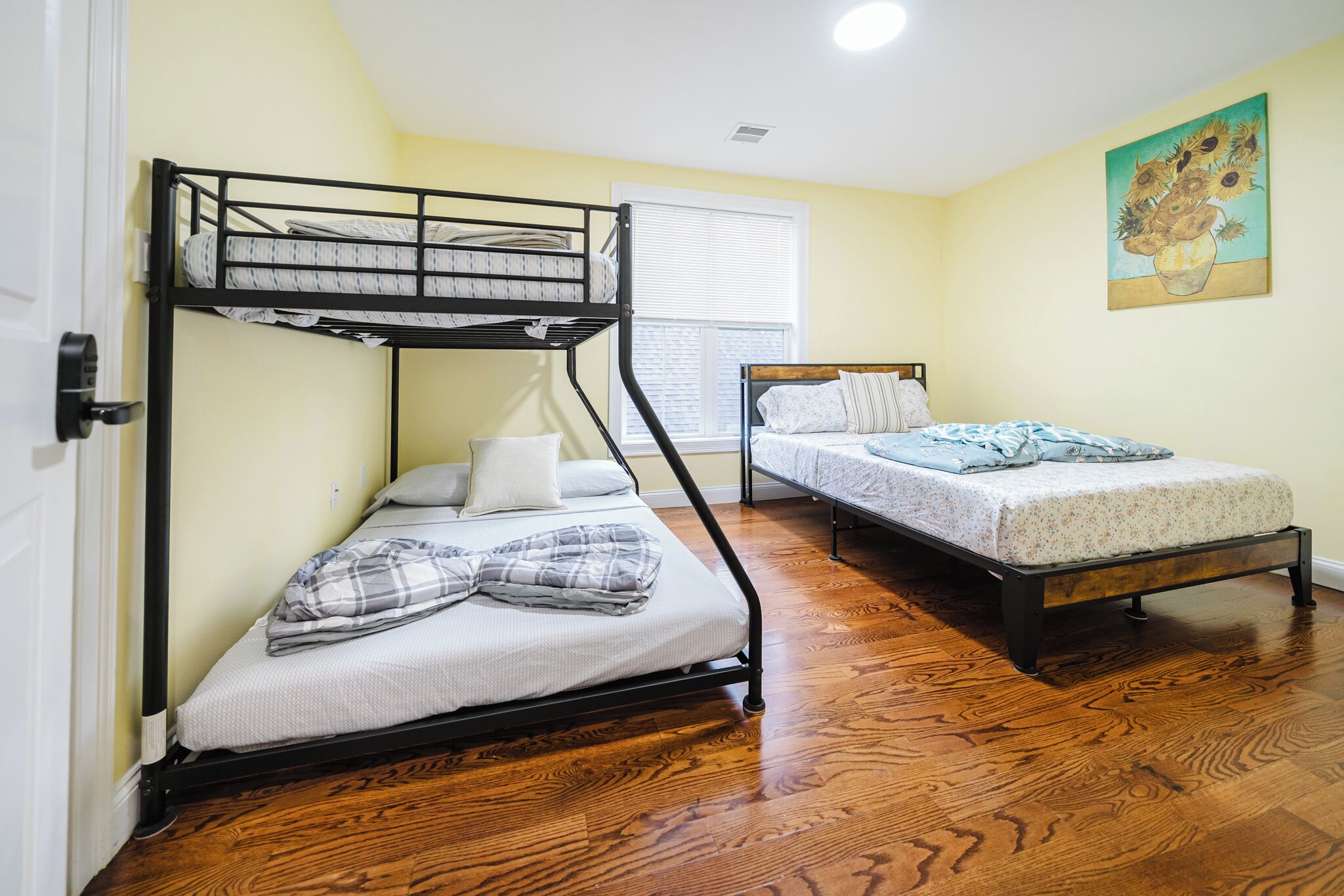 Una habitación con literas, una cama individual, suelo de madera, una ventana con persianas y un cuadro de girasol en la pared.