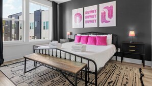 A modern bedroom with a large bed, a bedside table, a nightstand, and a window with a view of buildings. - NEW! Sleeps 20+ 2 SideBySide East Nashville Homes! (Nashville)
