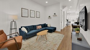 A modern living room with a blue sectional sofa, a small coffee table, and a floor lamp. There is a kitchen area visible in the background with white cabinets and a large mirror. - Eden Townhouse: Hot Tub & Golf Retreat w/Mountain Views (Eden)