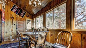 Interior de una cabaña de madera con una mesa de comedor, sillas y ventanas amplias que dan a un paisaje de árboles.