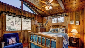 Un dormitorio rústico de madera con una cama, una silla, un ventilador de techo y una ventana con vistas a un paisaje nevado.