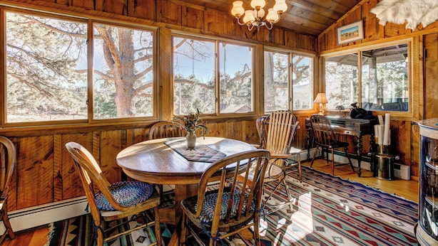 Interior de madera con una mesa redonda para comer, sillas, una araña y ventanas grandes que dan a vistas de árboles.