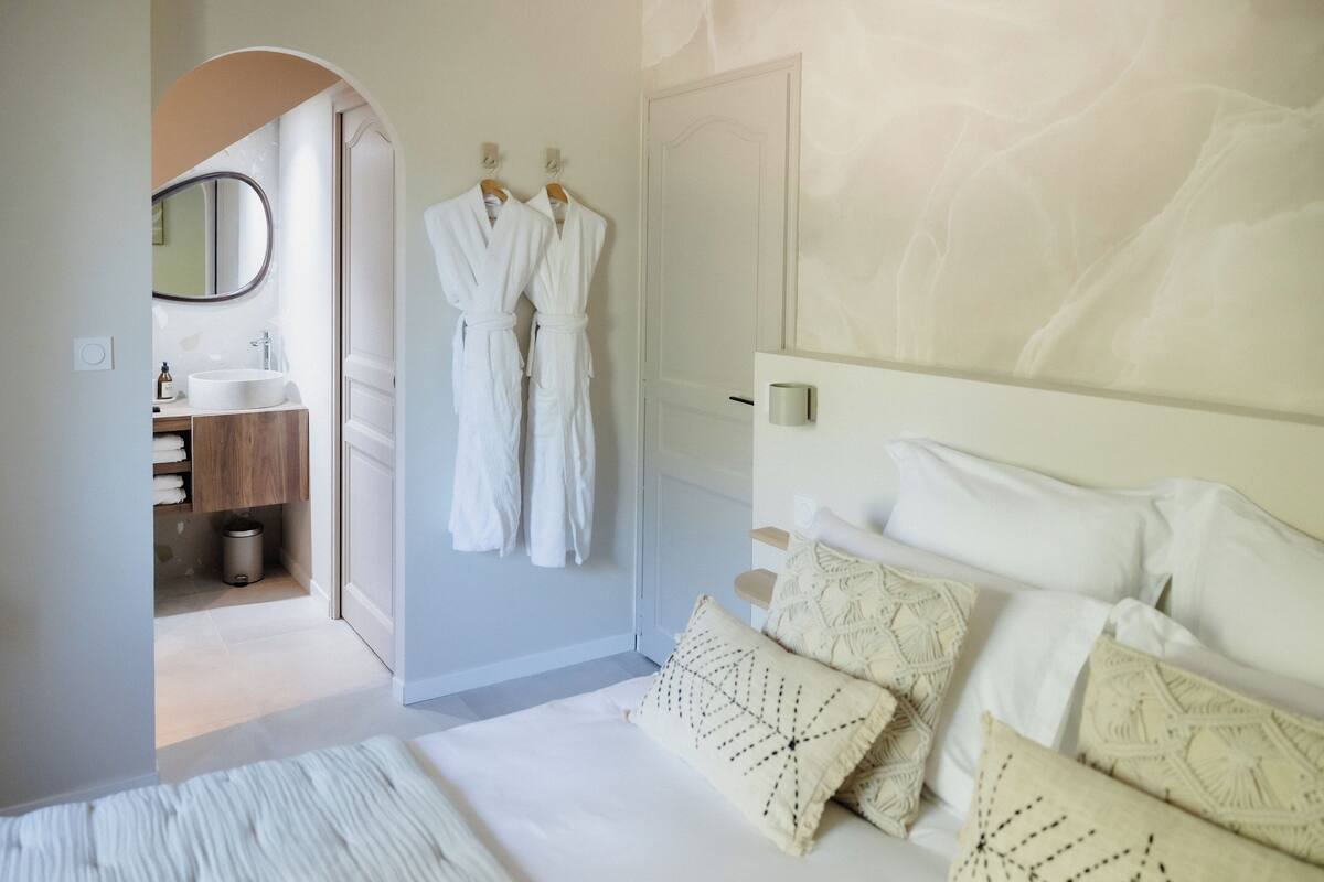 A bedroom with a bed, pillows, a bedside table, a mirror, and a door leading to a bathroom.