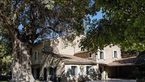 Un cortile con un grande albero, un edificio in pietra con tetto in tegole e una piccola costruzione anch'essa con tetto in tegole.