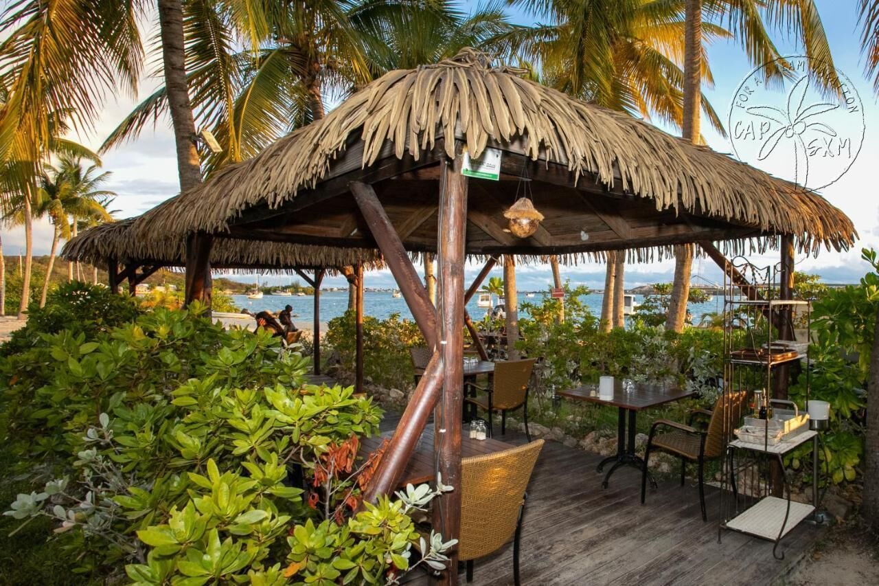 A thatched-roof outdoor seating area with tables and chairs, surrounded by lush greenery and palm trees, overlooking a beach.