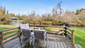 Eine Holzterrasse mit zwei Stühlen und einem Tisch, die auf einen Fluss mit Blick auf Bäume und ein Gebäude im Hintergrund blickt.