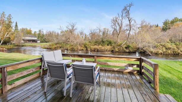 Una terraza de madera con dos sillas y una mesa, con vista a un río, rodeada de árboles y un edificio al fondo.