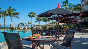 Un espace de restauration aménagé au bord de la piscine, avec des tables, des chaises, des parasols et des palmiers.